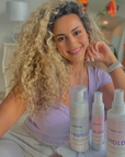 Model with curly blonde hair next to bottle of GLOSS Rose Meadow Hair Oil-InStyler