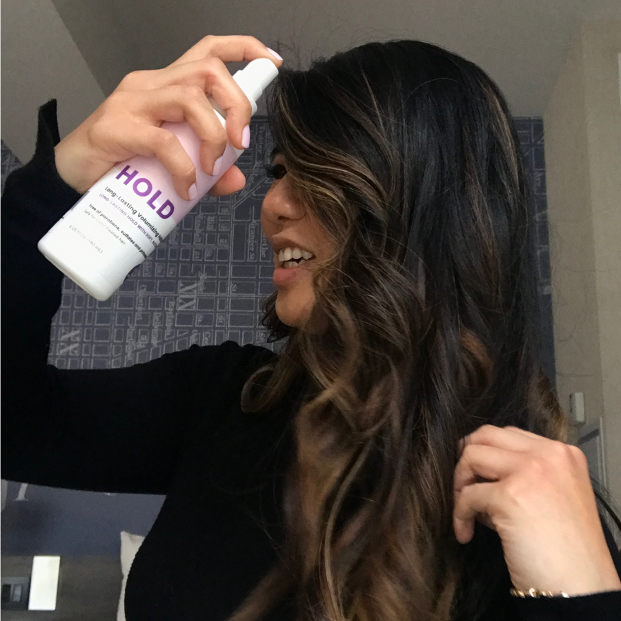 Photo of model with curly brown hair spraying her hair with HOLD Volumizing  Hair Spray-InStyler